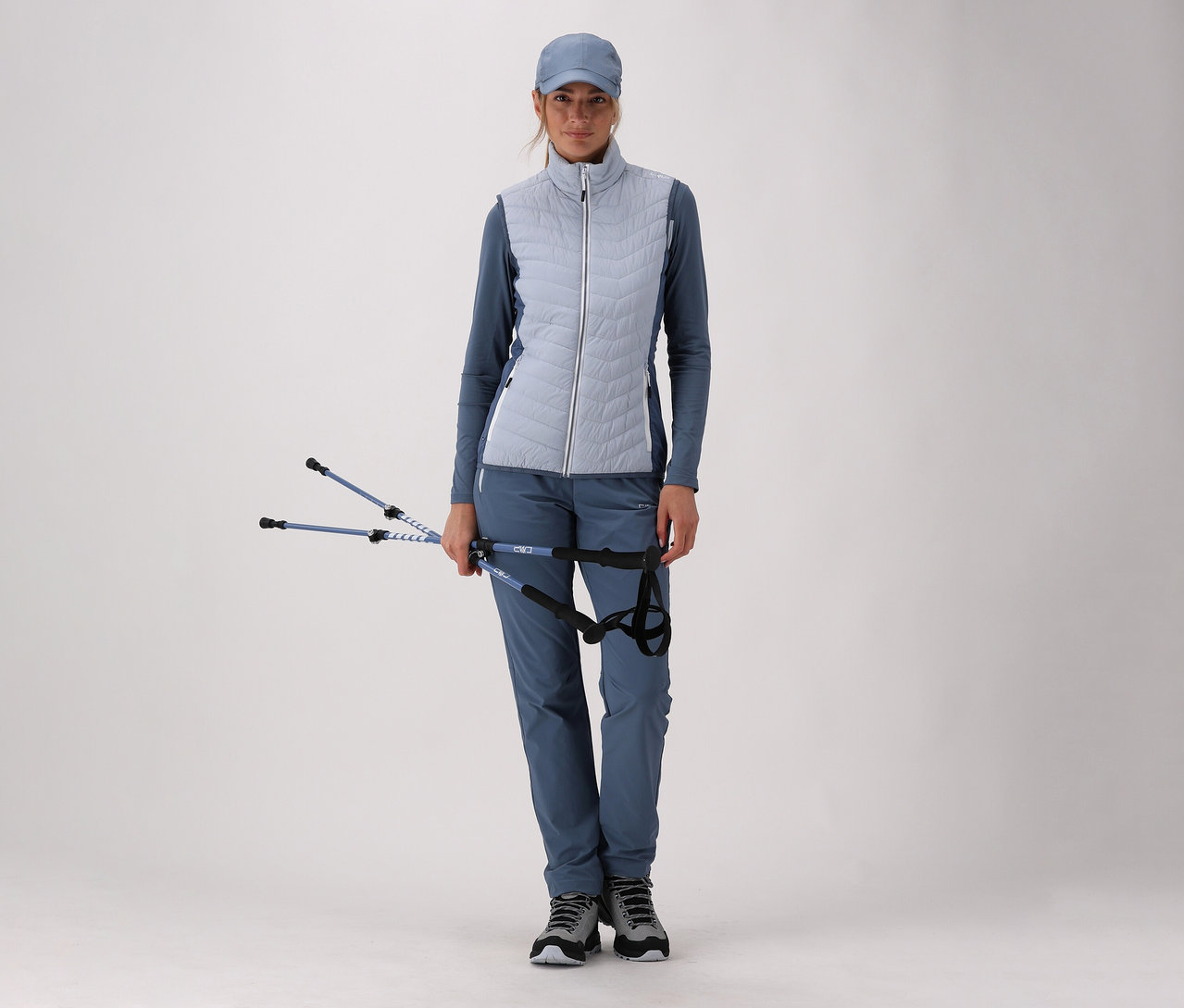 Femme en tenue de sport bleue avec une Gilet hybride CMP pour femme tenant des bâtons de randonnée sur fond clair.