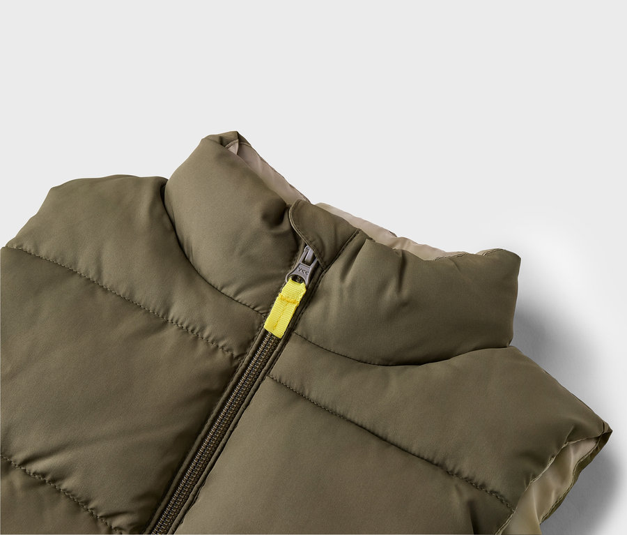 Gros plan d'un gilet matelassé vert olive avec fermeture éclair jaune et doublure beige.