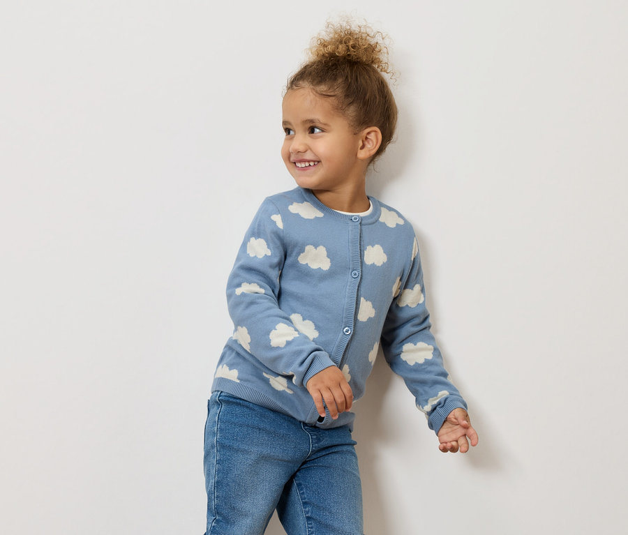 Une petite fille souriante aux cheveux bouclés porte un cardigan bleu à motifs de nuages et un jean.