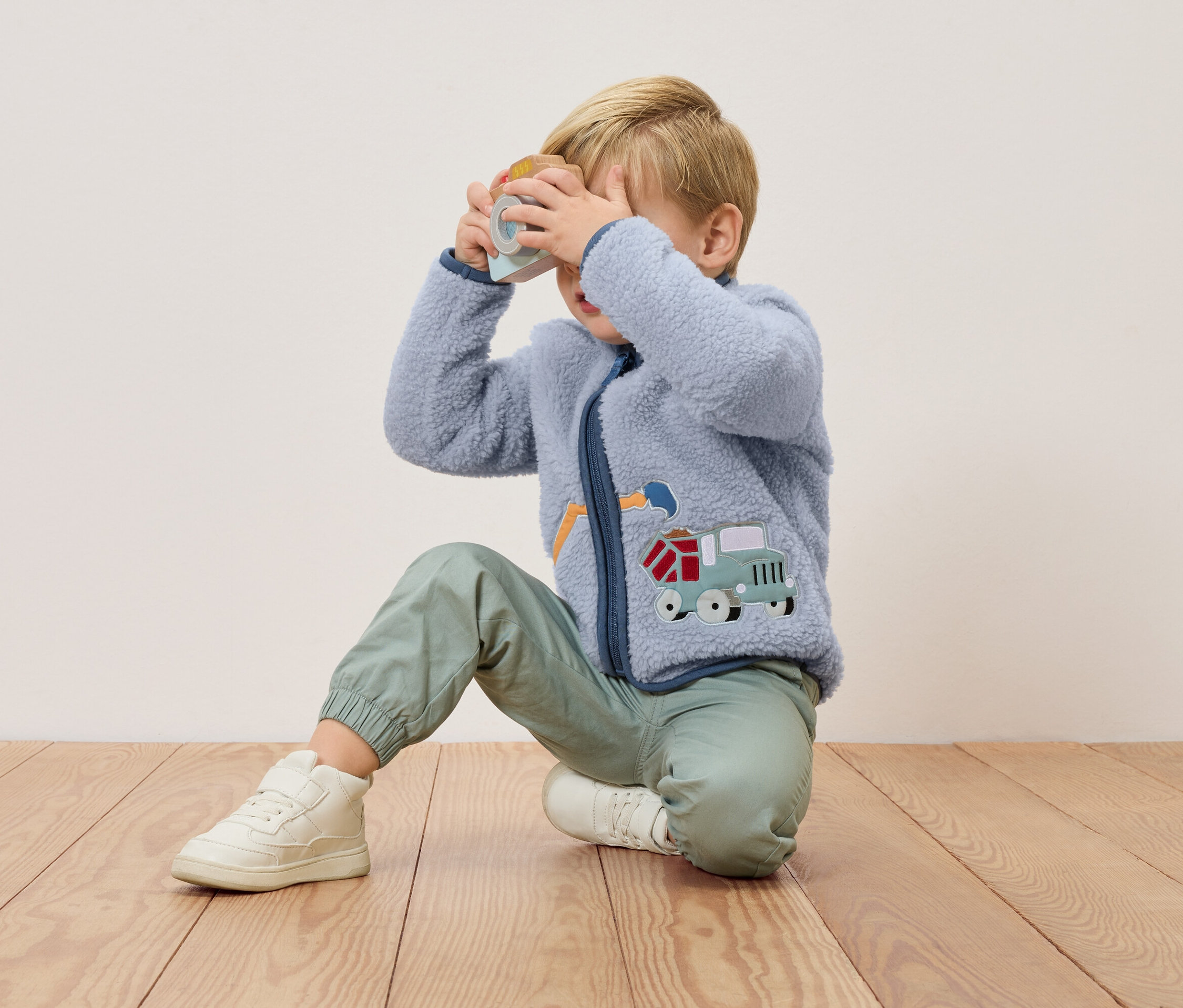 Un garçon blond est agenouillé sur un plancher en bois et tient un appareil photo jouet devant son visage. Il porte une veste en polaire bleue avec une application de camion et un pantalon vert.