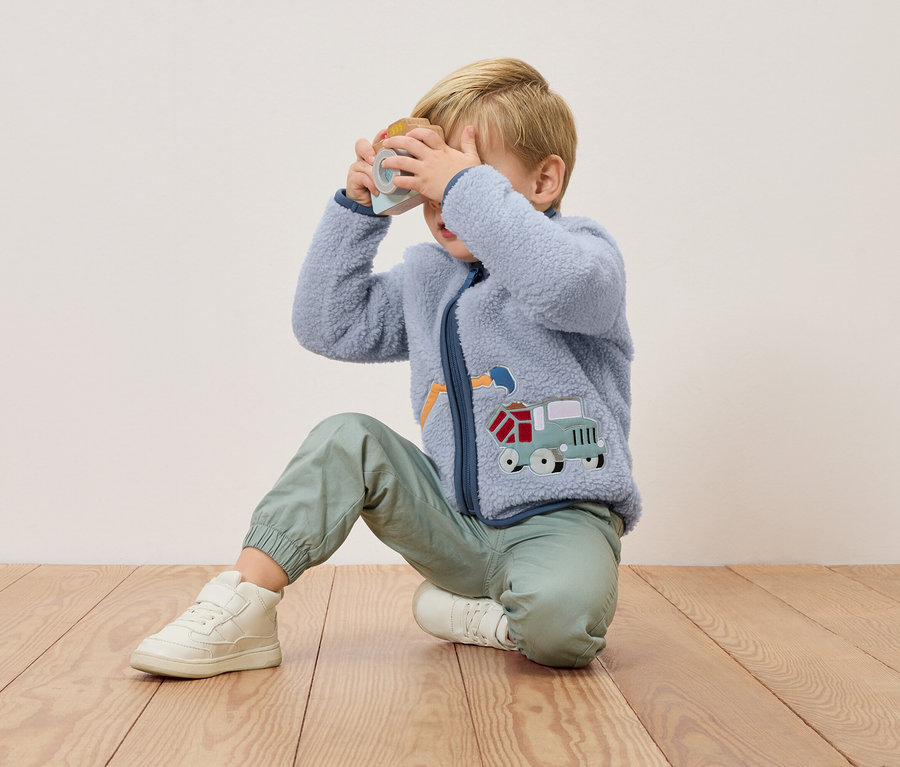 Un garçon blond est agenouillé sur un plancher en bois et tient un appareil photo jouet devant son visage. Il porte une veste en polaire bleue avec une application de camion et un pantalon vert.