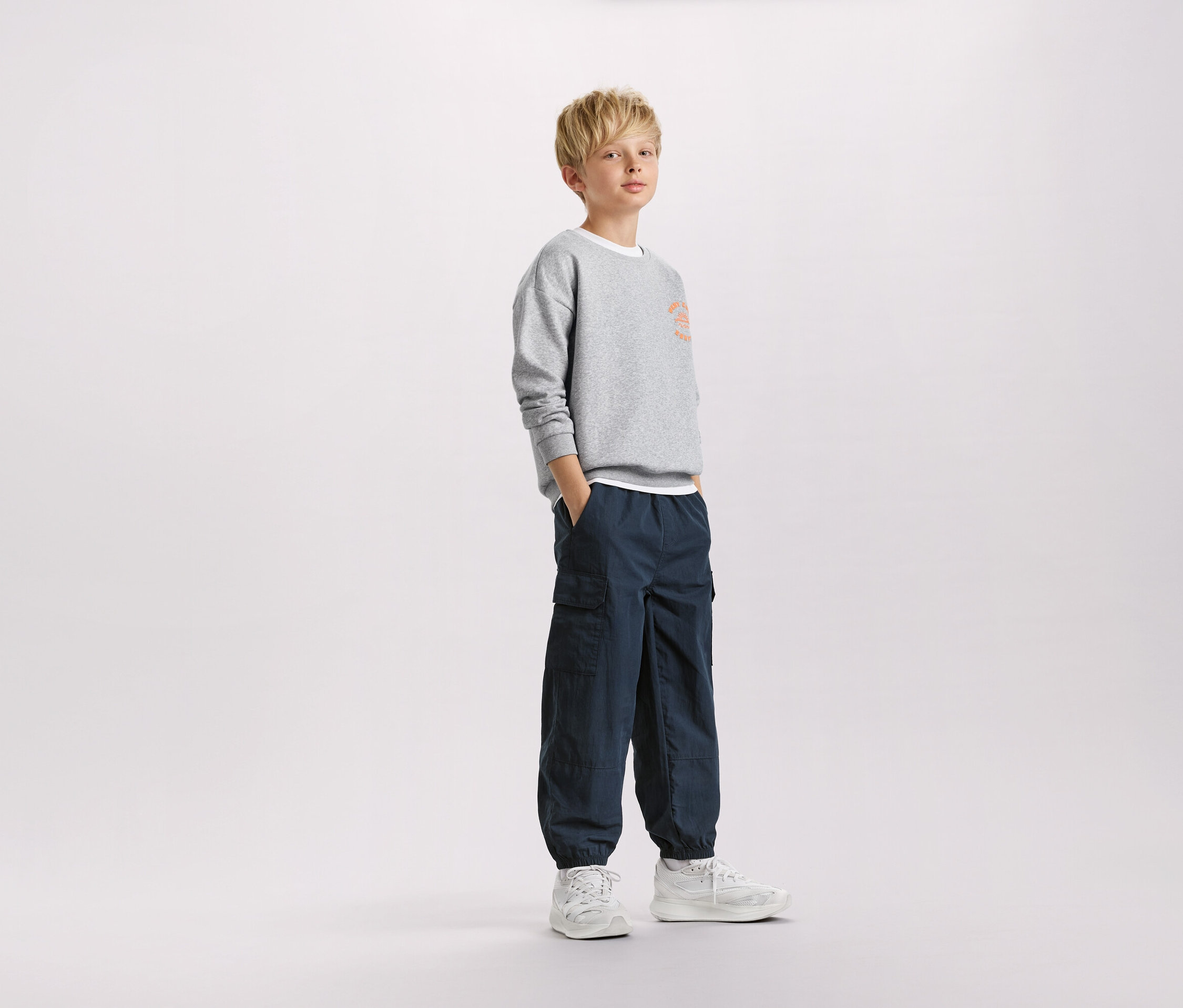 Un garçon blond se tient debout, les mains dans les poches, portant un sweat-shirt gris et un pantalon cargo bleu marine.