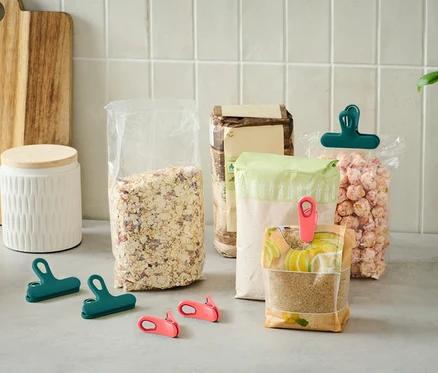 Divers emballages alimentaires ouverts sont posés sur un comptoir de cuisine, certains fermés par des pinces vertes et roses.