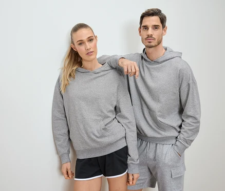 Une femme et un homme posent en sweat à capuche gris et short devant un mur blanc.