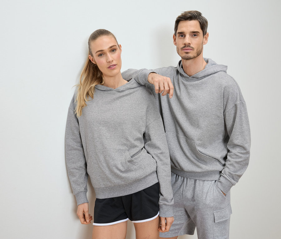 Une femme et un homme posent en sweat à capuche gris et short devant un mur blanc.