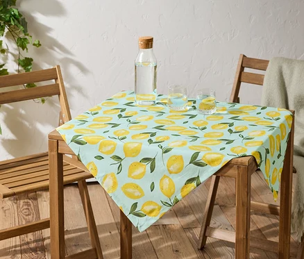 Une table avec une nappe bleu clair à motif de citron est placée entre deux chaises en bois. Une bouteille d'eau et deux verres d'eau sont posés sur la table.