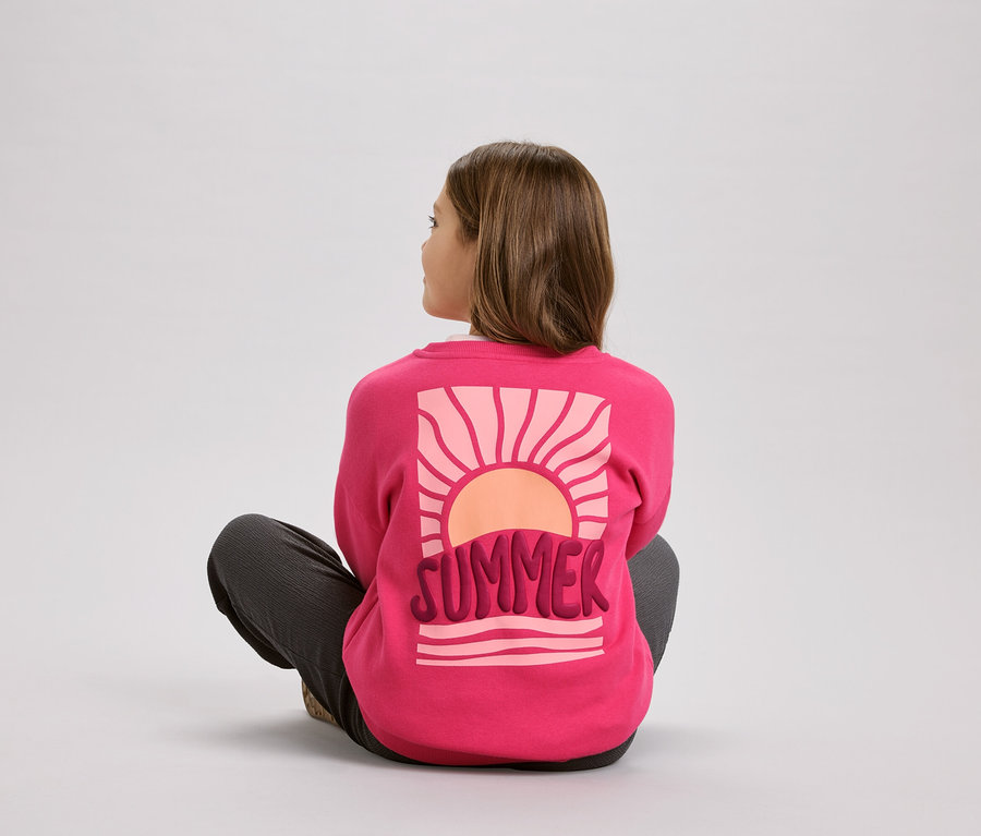 Une fille est assise en tailleur et porte un pull rose avec l'inscription « Summer » et un motif de soleil.