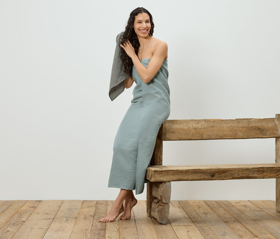 Une femme assise sur un banc en bois, enveloppée dans une serviette bleu clair, se sèche les cheveux avec une serviette grise.