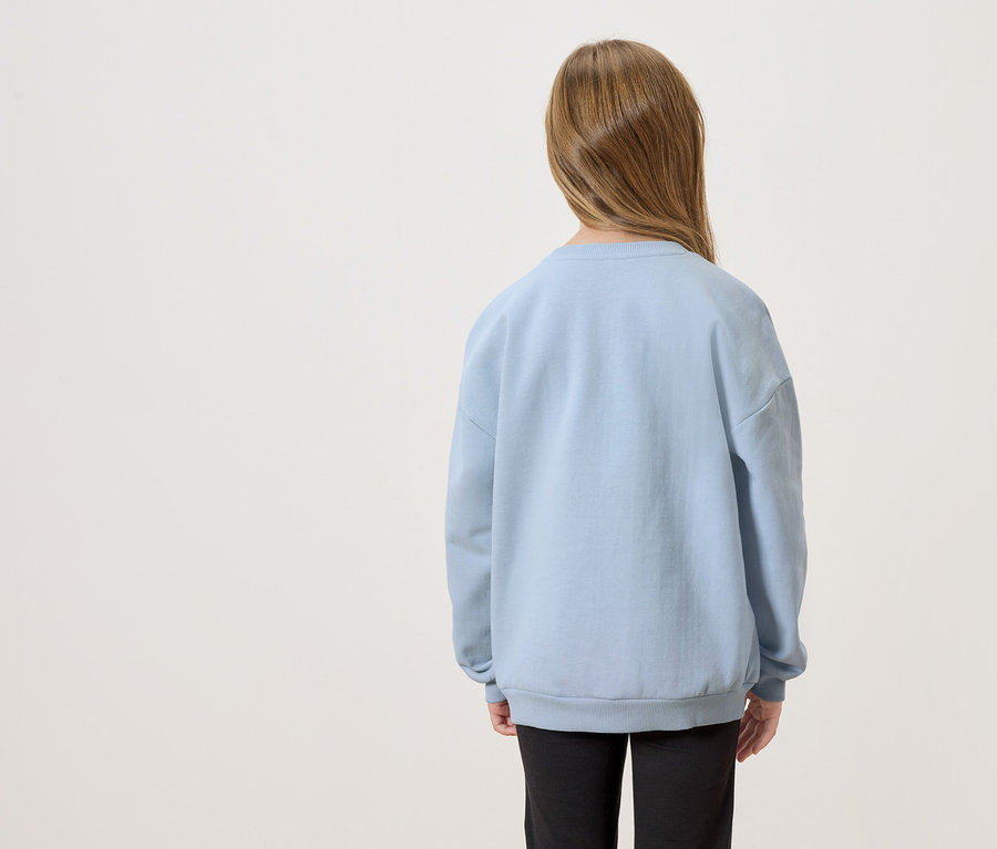 Vue de dos d'un enfant aux longs cheveux bruns portant un pull bleu clair et un pantalon noir.