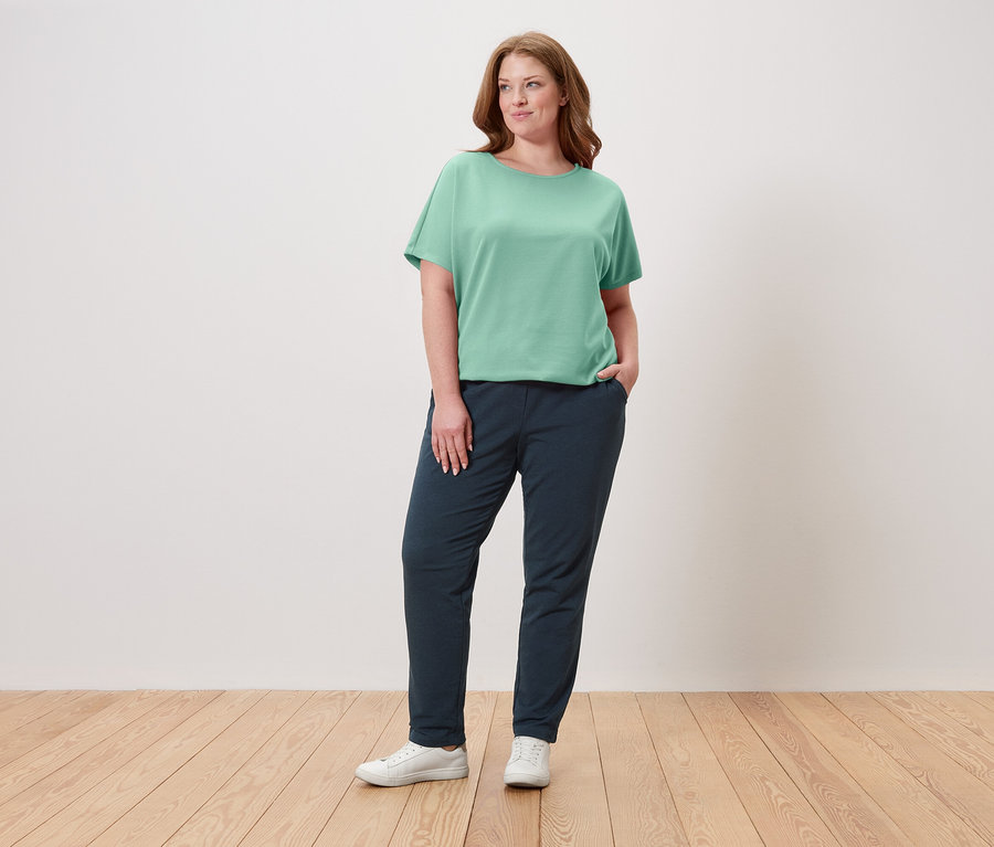 Une femme se tient debout, la main dans la poche, portant un t-shirt vert, un pantalon bleu marine et des baskets blanches.