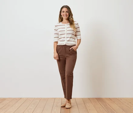 Une femme se tient devant un mur blanc, portant un cardigan rayé, un pantalon marron et des chaussures beiges.