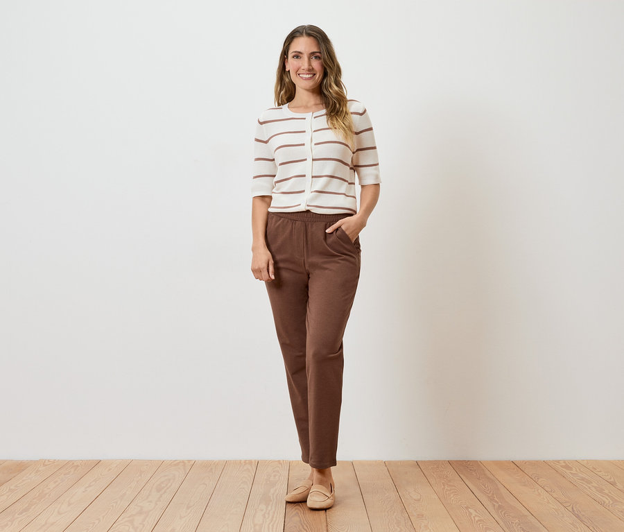 Une femme se tient devant un mur blanc, portant un cardigan rayé, un pantalon marron et des chaussures beiges.