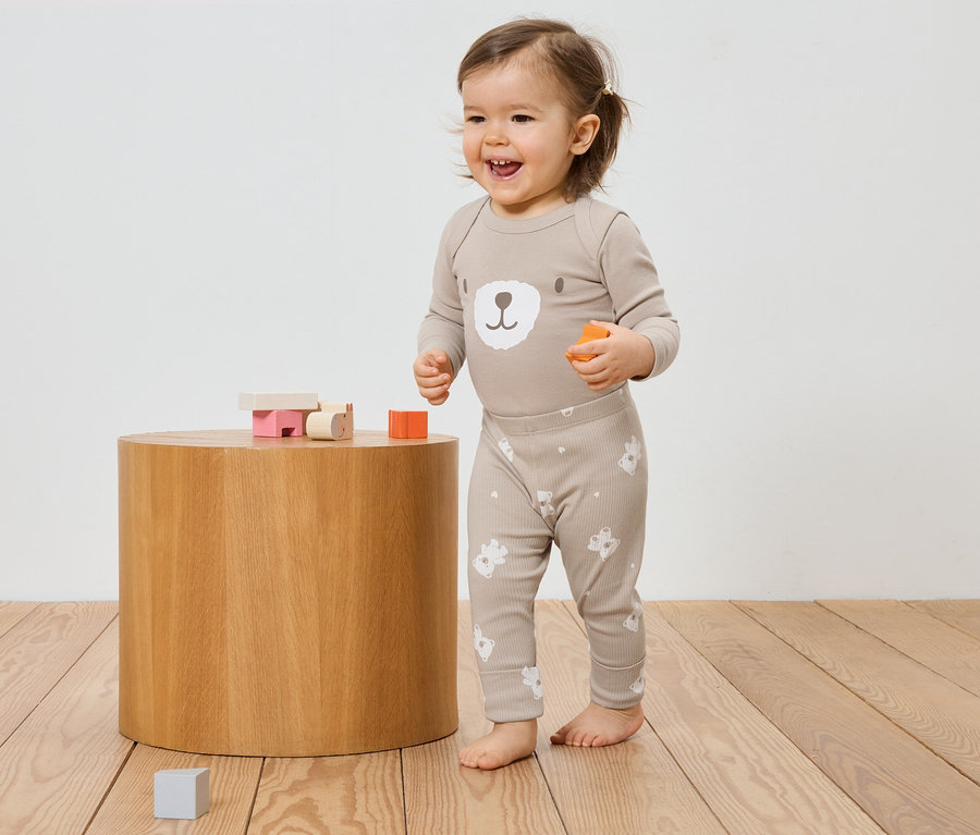 Un bébé souriant en pyjama beige à motif d'ours se tient à côté d'un tabouret en bois avec des jouets.