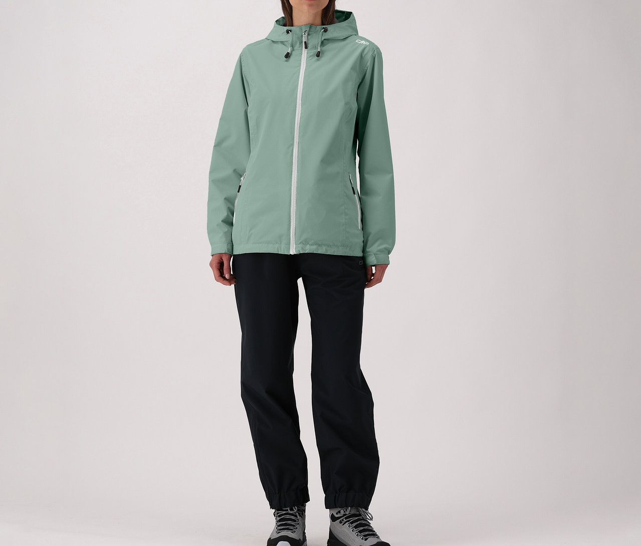 Femme portant une veste CMP vert menthe avec fermeture éclair blanche, un pantalon noir et des chaussures grises.