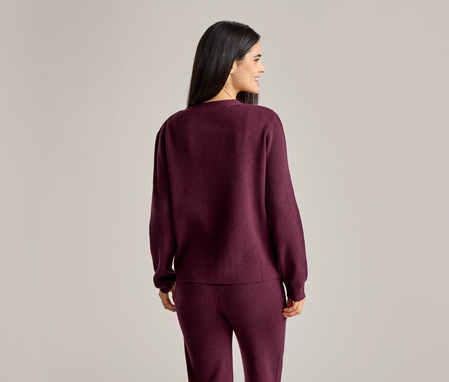 Femme vue de dos portant un pull et un pantalon côtelés bordeaux. Elle sourit et regarde vers la gauche.