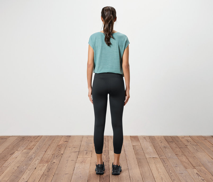 Vue de dos d'une femme avec une queue de cheval portant un haut bleu-vert, un legging noir et des baskets noires sur un plancher en bois.