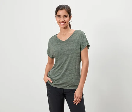 Femme brune souriante portant un t-shirt chiné vert olive et un pantalon foncé, une main dans la poche.