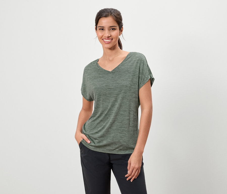 Femme brune souriante portant un t-shirt chiné vert olive et un pantalon foncé, une main dans la poche.