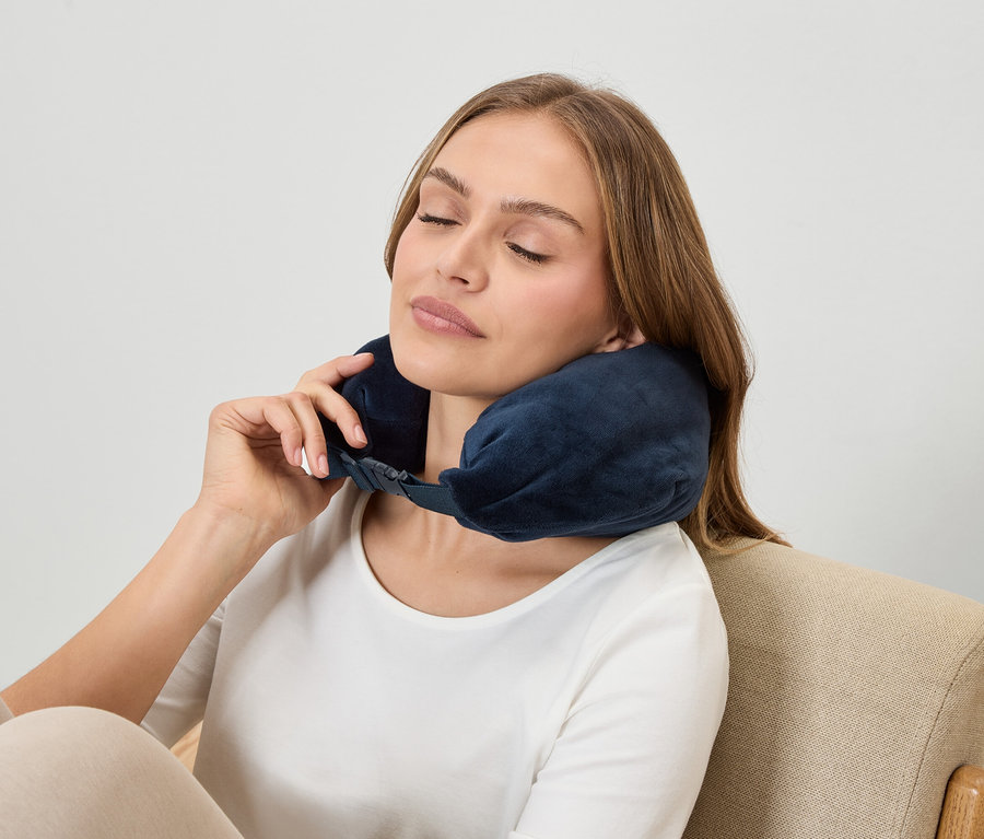 Une femme aux yeux fermés porte un oreiller cervical bleu marine tout en étant assise dans un fauteuil.