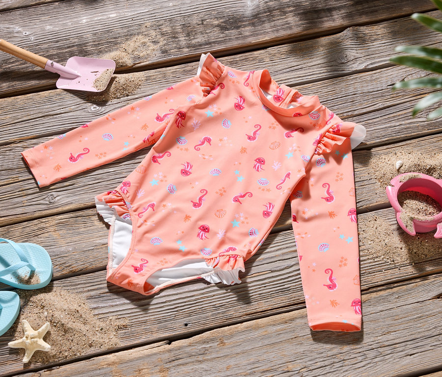 Un maillot de bain rose avec des motifs marins est posé sur une table en bois, accompagné d'une paire de tongs bleues, d'une pelle rose et d'un moule à sable rose.