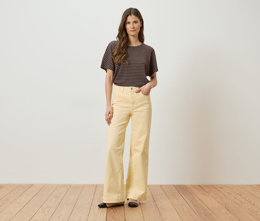 Une femme portant un t-shirt rayé et un pantalon évasé jaune, une main dans la poche, devant un mur blanc et un plancher en bois.