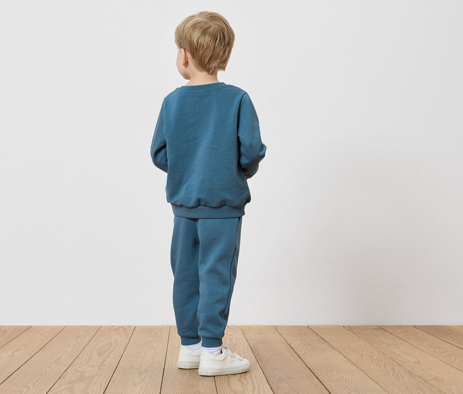 Un enfant aux cheveux blonds se tient dos au spectateur, portant un survêtement bleu marine et des baskets blanches.