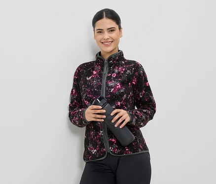 Une femme souriante en veste polaire à motifs tient une bouteille d'eau noire devant un mur blanc.