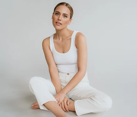 Une femme est assise pieds nus en tailleur sur le sol, vêtue d'un débardeur blanc et d'un jean blanc Angels Denim « Evy Cool » O-Shape.
