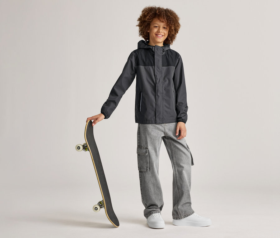 Un garçon souriant aux cheveux bouclés se tient debout avec une planche à roulettes à la main, portant une veste gris et noir, un jean gris et des chaussures blanches.