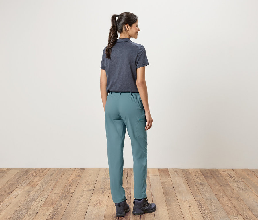 Vue de dos d'une femme avec une queue de cheval portant un polo bleu marine, un pantalon vert et des chaussures de randonnée devant un mur blanc et un plancher en bois.