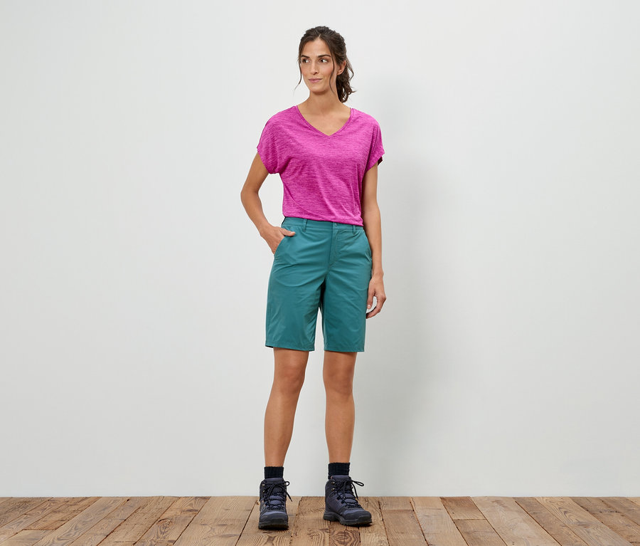 Une femme se tient debout, la main dans la poche, portant un t-shirt rose, un short vert et des chaussures de randonnée.