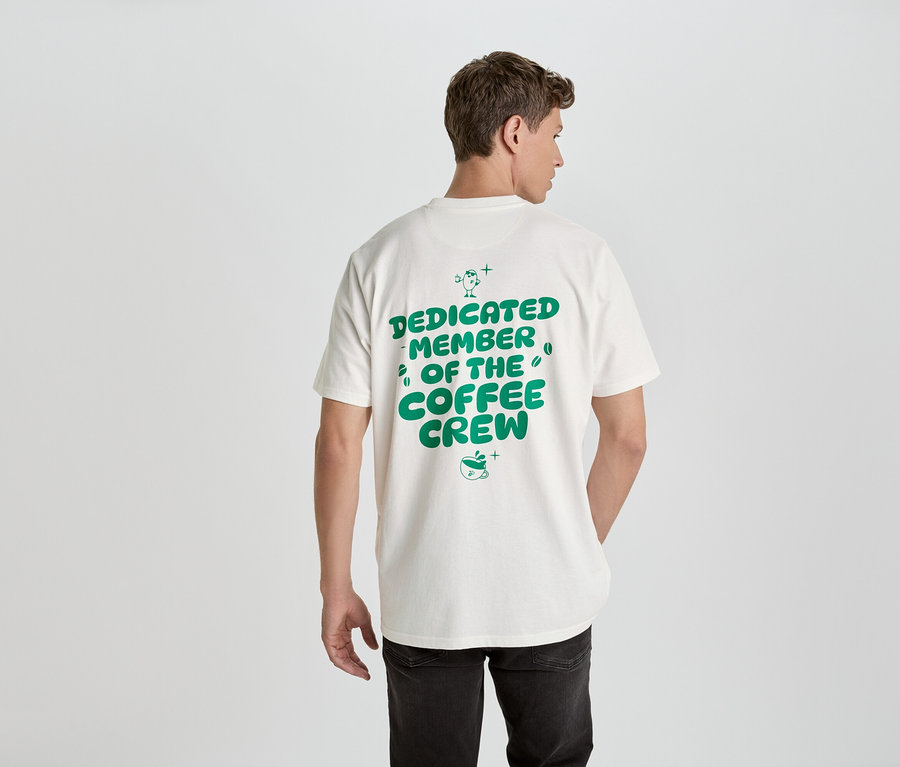 Un homme vu de dos porte un t-shirt blanc avec l'inscription verte 'Dedicated Member of the Coffee Crew'.