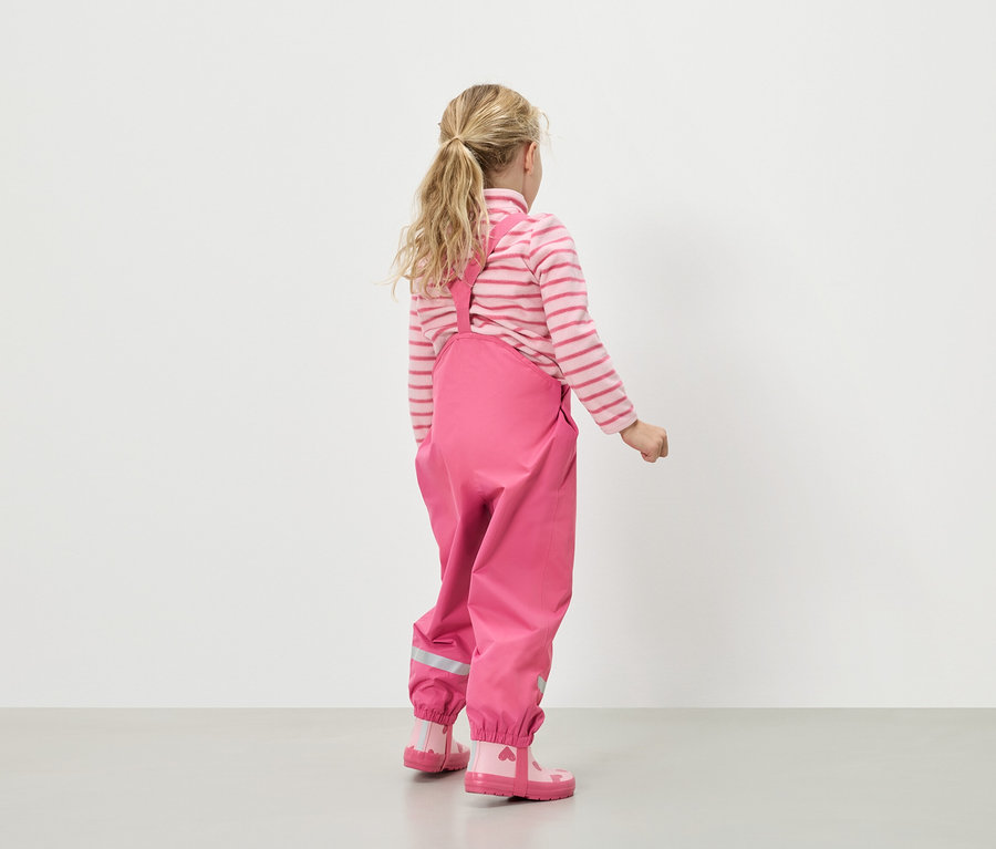 Vue de dos d'une fillette blonde portant un pull rayé rose et blanc, un pantalon de pluie rose à bretelles et des bottes en caoutchouc roses à cœurs.