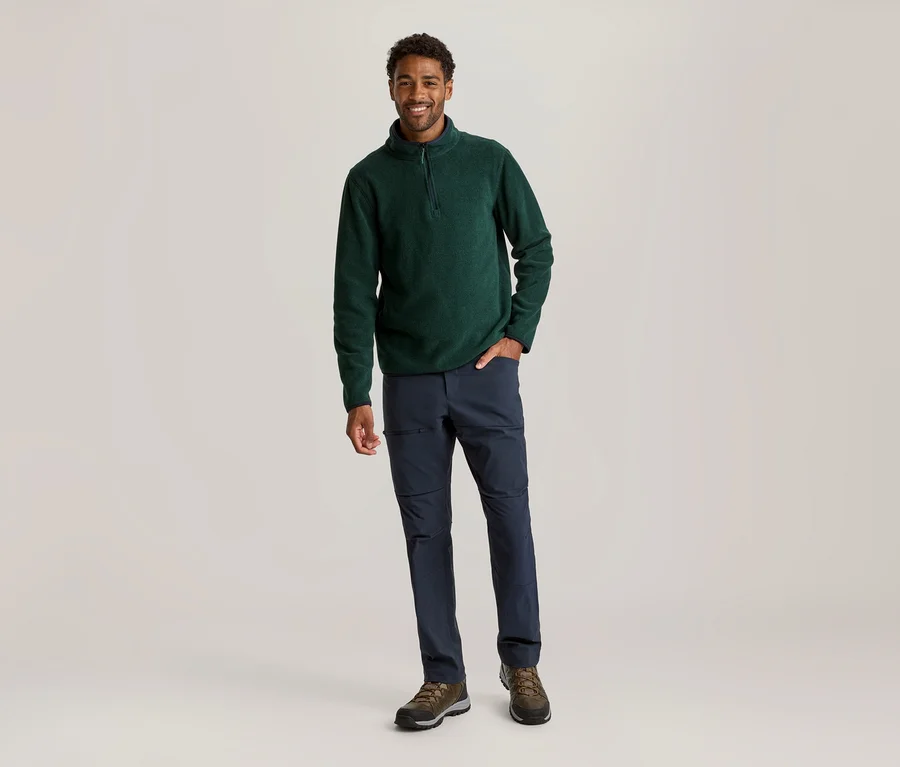 Un homme souriant est debout, portant un pull polaire vert foncé à demi-zip, un pantalon bleu foncé et des chaussures de randonnée marron. Il a la main droite dans la poche de son pantalon.