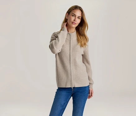 Femme aux cheveux longs et châtains clairs, portant un cardigan zippé côtelé beige et un jean bleu. Elle touche sa nuque.