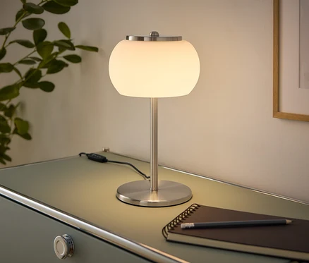 Une lampe de table allumée avec un abat-jour sphérique blanc et une base en métal brossé est posée sur une commode vert clair. À côté se trouvent un carnet et un crayon. Une plante est à gauche, un tableau encadré est au mur à droite.
