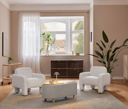 Un salon confortable avec deux fauteuils gris clair et un pouf assorti sur un tapis. Une lampe de table à LED sans fil beige est posée sur une armoire en bois clair à côté d'une grande plante. Un meuble de rangement marron foncé se trouve sous une fenêtre en baie.
