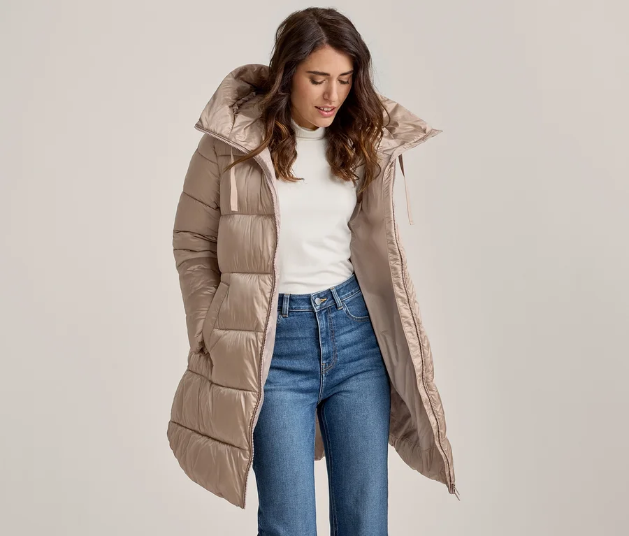 Femme aux longs cheveux bruns portant un long manteau matelassé beige ouvert sur un col roulé blanc et un jean bleu, regardant vers le bas.