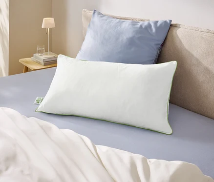 Un lit avec un drap bleu clair, une couette blanche et deux oreillers. Un oreiller blanc à bordure verte est posé devant un oreiller bleu clair. À gauche, une table de chevet avec une lampe et un verre d'eau.