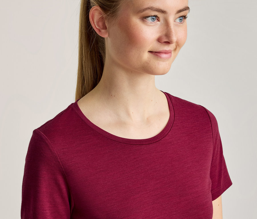 Jeune femme aux cheveux blonds attachés en queue de cheval, portant un t-shirt bordeaux à col rond, souriant et regardant vers la droite.