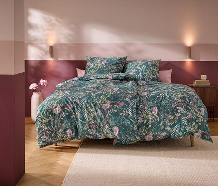 Un lit avec une housse de couette verte à motifs floraux et deux oreillers dans une chambre aux murs rayés horizontalement roses et bordeaux. Deux appliques murales éclairent le mur au-dessus du lit. Un vase blanc avec des fleurs roses est à gauche, et une table de chevet en bois avec des livres et une bougie est à droite. Un tapis clair recouvre une partie du parquet.