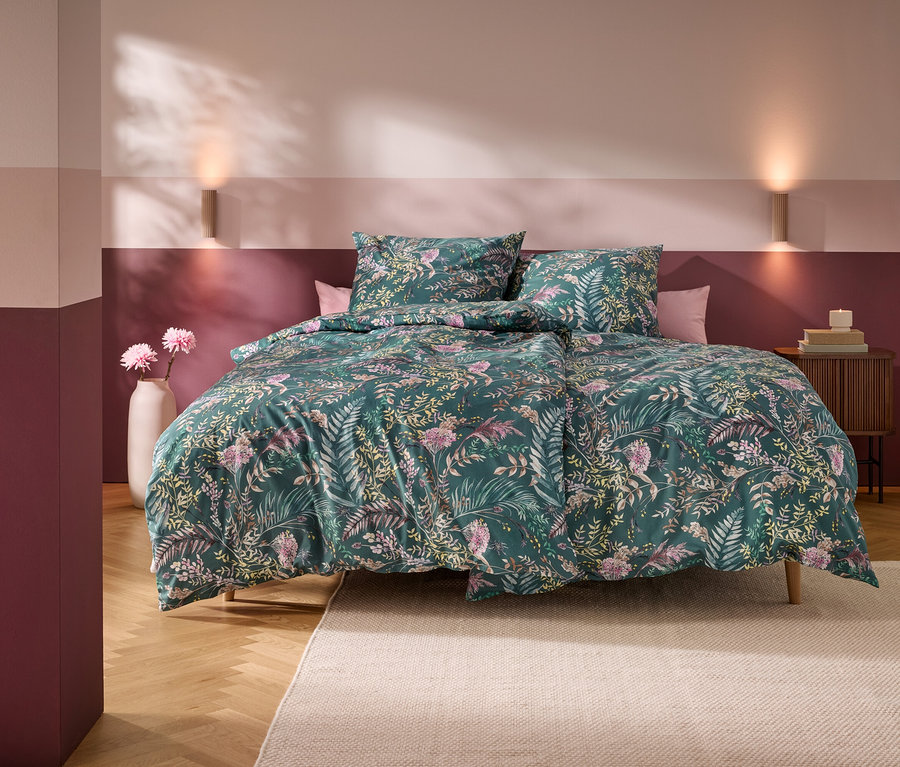 Un lit avec une housse de couette verte à motifs floraux et deux oreillers dans une chambre aux murs rayés horizontalement roses et bordeaux. Deux appliques murales éclairent le mur au-dessus du lit. Un vase blanc avec des fleurs roses est à gauche, et une table de chevet en bois avec des livres et une bougie est à droite. Un tapis clair recouvre une partie du parquet.