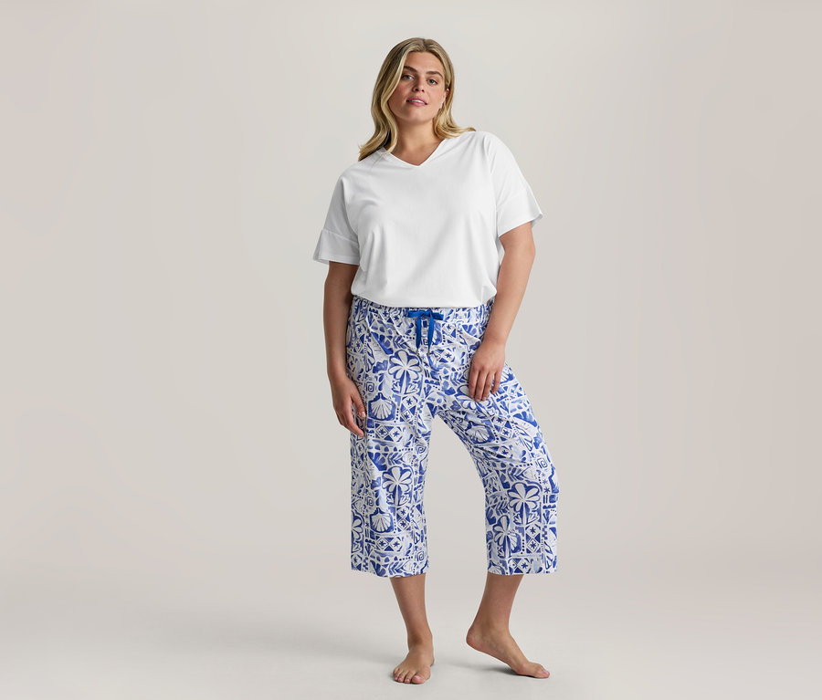 Femme debout, portant un t-shirt blanc à col en V et un pantalon de 3/4-Pyjama-Set à motifs bleus et blancs avec un cordon de serrage bleu, pieds nus.