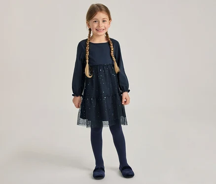 Jeune fille souriante portant une robe bleu marine à manches longues avec une jupe en tulle pailleté, des collants bleu marine et des chaussures en velours. Elle a deux tresses.