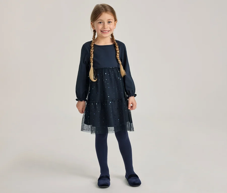 Jeune fille souriante portant une robe bleu marine à manches longues avec une jupe en tulle pailleté, des collants bleu marine et des chaussures en velours. Elle a deux tresses.