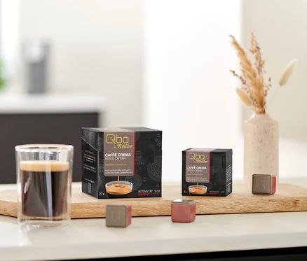 Une planche en bois sur un comptoir de cuisine blanc avec un verre de café, des biscuits, deux paquets de Qbo Caffè MENOS CAFEÍNA (27 et 8 capsules) et deux capsules individuelles. À droite, un vase avec de l'herbe séchée.