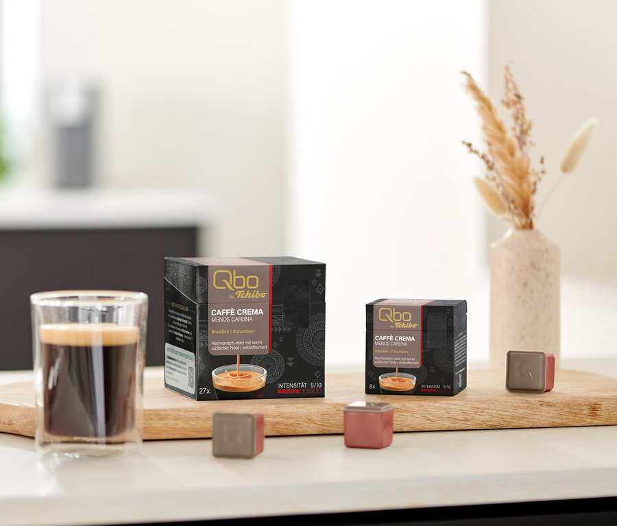 Sur une table, des paquets de capsules Qbo Caffè MENOS CAFEÍNA et une tasse de café sur une planche en bois.