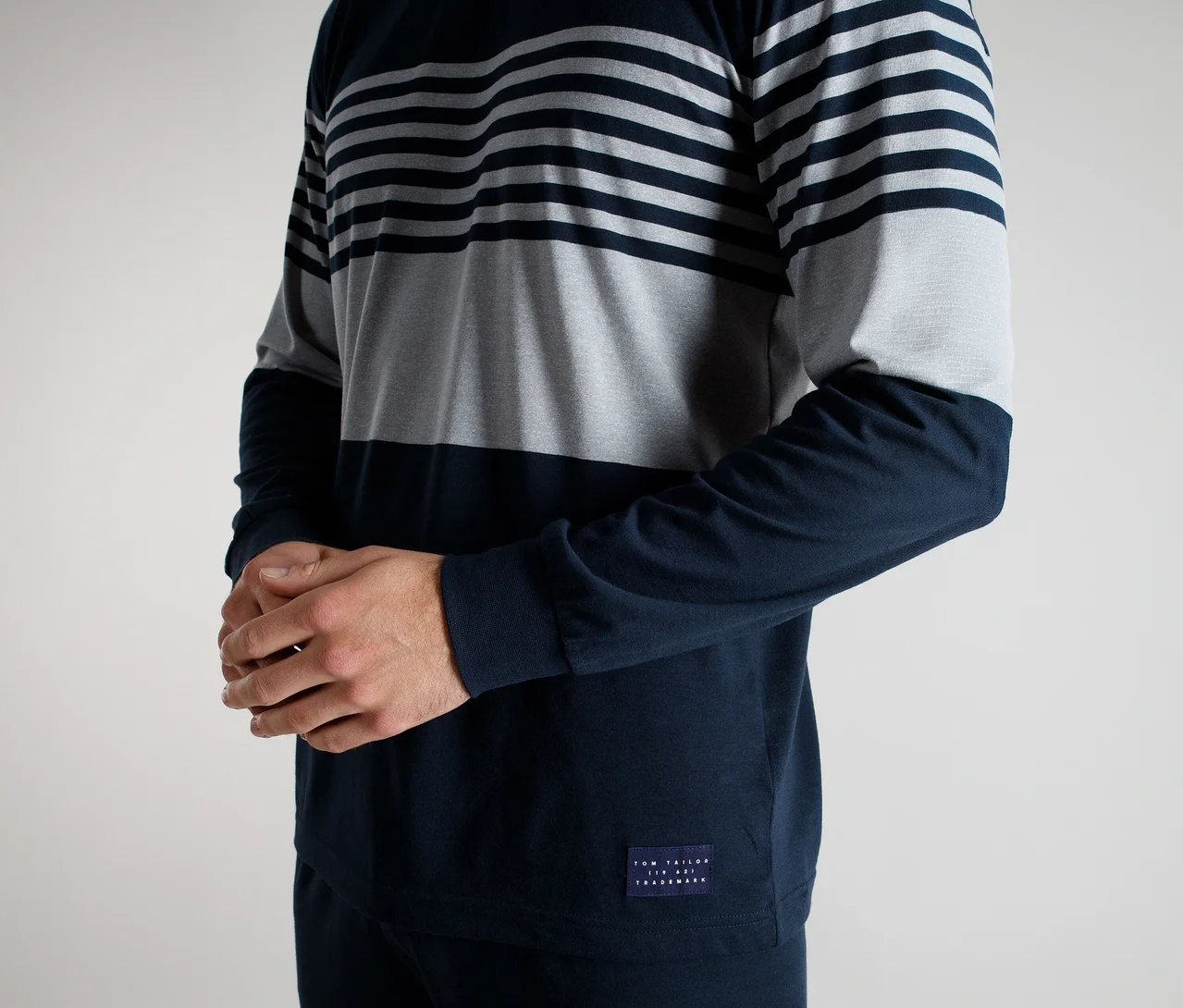 Homme portant un t-shirt à manches longues avec des rayures horizontales bleu foncé et grises en haut, une partie centrale grise unie et un bas et des manches bleu foncé unis. Ses mains sont jointes.