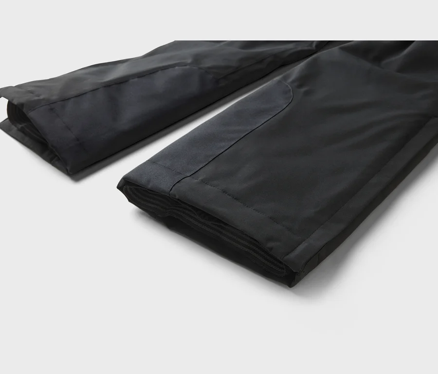 Deux jambes de pantalon noires pliées sur un fond gris clair, montrant un tissu lisse, des empiècements renforcés et des poignets intérieurs côtelés.
