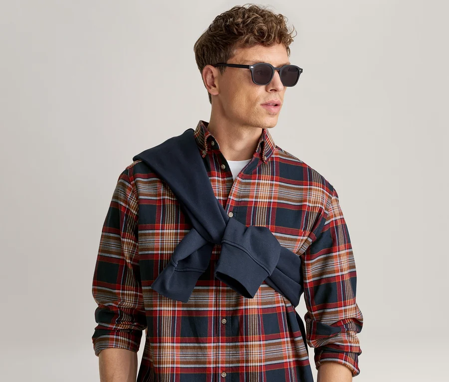 Homme en chemise à carreaux rouge, bleue et blanche, lunettes de soleil noires et sweat-shirt bleu marine noué sur les épaules.